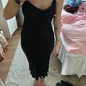 Svart långklänning i satin - Svart långklänning med draperad urringning och smala axelband. Klänningen har en figurnära passform och faller snyggt längs kroppen. 