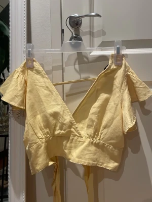 Gul omlottopp med knytband i storlek S från Bik Bok - Söt ljusgul omlottopp med korta, fladdriga ärmar och djup v-ringning. Toppen har breda knytband i midjan som knyts bak och ger en croppad look. Tillverkad i ett lätt bomullsliknande material, perfekt för varma dagar.