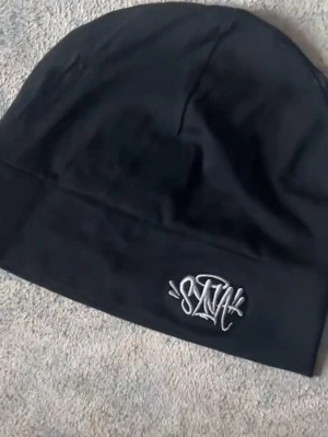 Svart mössa med broderad logga - Svart mössa i mjukt tyg med broderad vit SNAK-logga framtill. Enkel och stilren design som passar till streetwear och vardagsoutfits. Mössan har en klassisk form och är lätt att matcha med olika stilar.