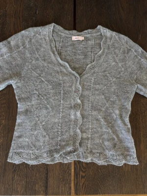 Grå kofta med volangkant från Nelly S - Söt gråmelerad kofta från Melly med volangkant längs nederkant och ärmslut. Koftan har v-ringning, knappar framtill och ett tunt, stickat material som ger en luftig känsla. Perfekt att slänga över en topp för en chill look.