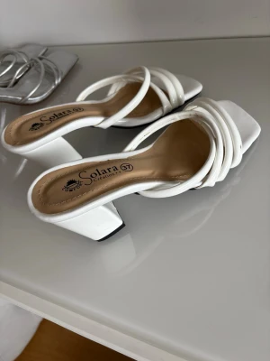 Vita ballerinaskor från Solara - Snygga vita klackskor från Solara med fyrkantig tå och bred klack. Skorna har flera tunna remmar över foten och en bekväm innersula i beige. Perfekta för dig som vill ha en trendig och stilren look.