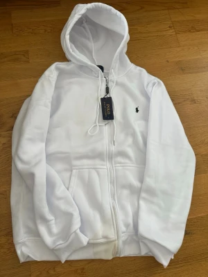 Vit hoodie från Polo Ralph Lauren - Snygg vit hoodie från Polo Ralph Lauren med dragkedja framtill och klassisk huva med dragsko. Broderad logga i svart på bröstet och två stora fickor framtill. Tillverkad i mjukt material som känns skönt mot huden. Perfekt för en clean och stilren look.
