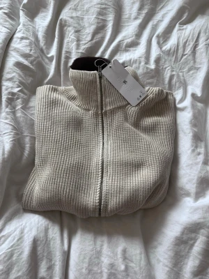 Beige ribbad ziptröja från Treton UF (Pris diskuterbart) - Säljer en beige ribbad ziptröja från Treton med hög krage och brun fuskpäls på insidan av kragen. Tröjan har dragkedja framtill och är tillverkad i ett mjukt stickat material. Perfekt för dig som gillar stilrena och mysiga plagg.