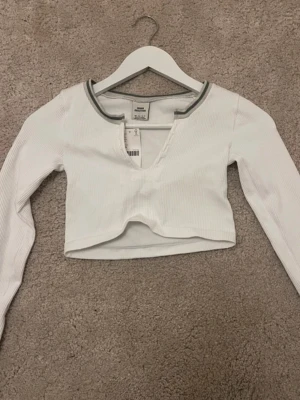 Vit croppad topp från Monki - Säljer en vit croppad topp från urbanoutfitters  med lång ärm och ribbad struktur. Toppen har en djup v-ringning och en grå detalj runt halsen som ger en sportig vibe. Perfekt att styla med jeans eller kjol. Kostar egentligen 400kr 
