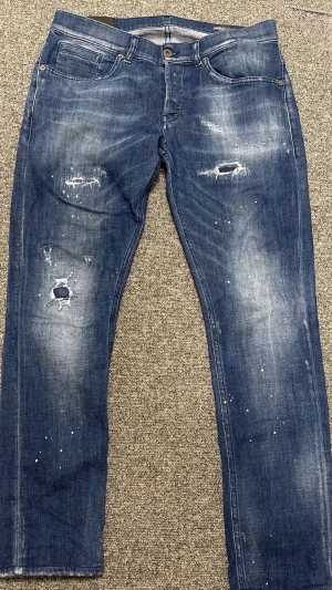 Mörkblå dondup george  - Säljer ett par blå dondup george med slitningar, hål och färgstänk för en trendig och avslappnad look. Jeansen har en klassisk femficksdesign. Perfekta för dig som vill klä upp dig men ändå se avslappnad ut och vill ha ett par unika jeans i garderoben. Skriv vid fler bilder och minsta lilla instrsse!