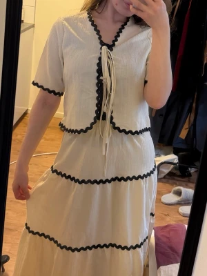 Vit set (kjol & topp) med svarta detaljer - Vit set i luftig bomull med svarta vågiga band längs kanterna och volanger. Toppen har korta ärmar, v-ringning och knytband framtill. Perfekt för en somrig look med unik design.