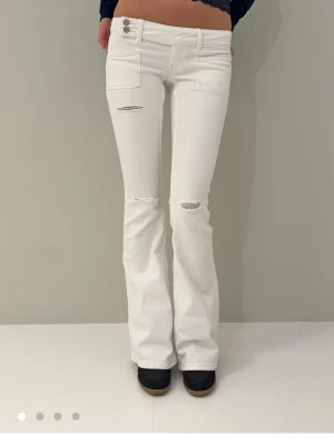Vita bootcut jeans med slitningar - Säljer ett par vita bootcut jeans med slitningar vid knäna och framfickor. Jeansen har hög midja, dubbla knappar och utsvängda ben. Perfekta för en trendig och avslappnad look. Har använt 1 gång och ser helt nya ut!