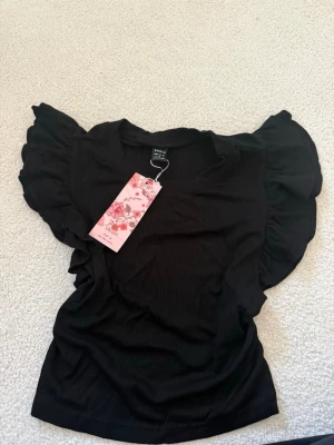 Svart volangtopp från SHEIN - Trendig svart topp från SHEIN med volangärmar och rund halsringning. Toppen är ribbad och har en figurnära passform som ger en snygg siluett. Perfekt för dig som gillar en stilren men ändå lekfull look.