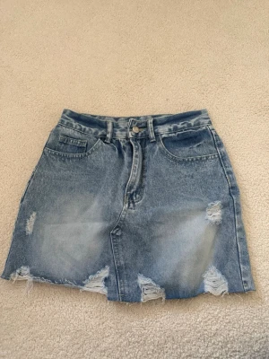 Blå jeanskjol med slitningar - Säljer en blå jeanskjol med rå kant och slitna detaljer framtill. Kjolen har klassisk femficksdesign, knapp och dragkedja i midjan samt bälteshällor. Perfekt för en avslappnad och trendig look.