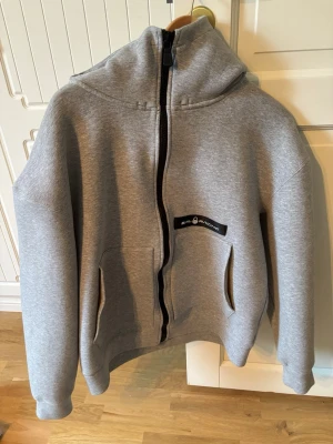 Grå zip hoodie från Sail Racing - Säljer en stilren grå hoodie från Sail Racing med dragkedja framtill och två fickor. Hoodien har en klassisk huva och en svart logopatch på bröstet. Perfekt för dig som gillar enkel och sportig stil. Materialet känns mjukt och bekvämt.