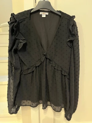 Svart blus med volanger från Gina Tricot - Säljer en svart blus från Gina Tricot med transparenta långärmar och små prickar i tyget. Blusen har volangdetaljer längs bröstet och axlarna samt en djup V-ringning. Perfekt för dig som gillar trendiga detaljer och en luftig känsla.