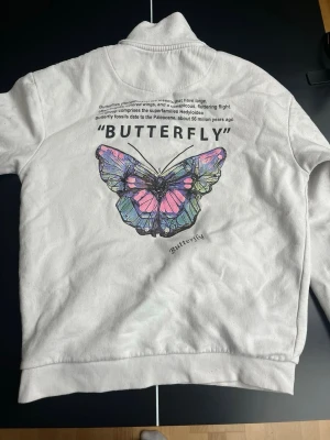 Vit halvzip tröja med fjärilstryck - Vit långärmad halvzip tröja med stor färgglad fjäril på ryggen och texten 'BUTTERFLY'. Mindre fjärilsmotiv och texten 'APPROACH' på bröstet. Tröjan har ribbade muddar och hög krage med dragkedja. Perfekt för dig som gillar streetwear och coola prints. Lite fläckig som man ser på sista bilden