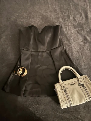 Svart axelbandslös kort klänning - Snygg svart axelbandslös kort klänning med hjärtformad urringning och lätt utsvängd kjol. Klänningen har en elegant, slät yta och är perfekt för dig som vill ha en stilren look. Materialet känns mjukt och har en lätt glans.