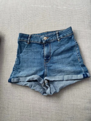 Jeansshorts med högmidja  - Ett par klassiska blåa jeansshorts med hög midja och uppvikta benslut. Shortsen har bälteshällor, knapp och dragkedja framtill samt två bakfickor. Mycket bra skick. Storlek: 34