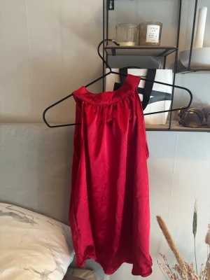 Röd axelbandslös sidenlinnetopp - Säljer en röd axelbandslös linnetopp i siden-look med loose passform. Toppen har en enkel, elegant design med rynkad kant upptill och är perfekt för dig som gillar stilrena plagg med lite extra lyster.