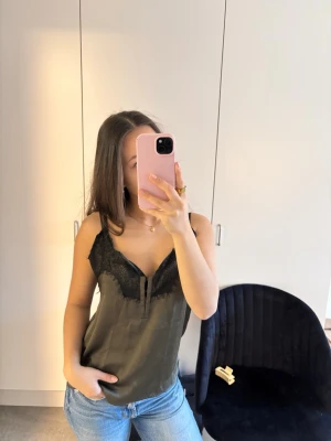 Olivgrön linnetopp med spetsdetaljer - Snygg olivgrön linnetopp med tunna axelband och svart spets längs urringningen. Toppen har en v-ringad form och är tillverkad i ett glansigt material som ger en lyxig känsla. Perfekt att styla med jeans eller kjol för en trendig look.