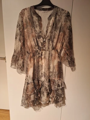 Kort klänning med ormmönster och volang - Säljer en kort klänning med ormmönster i beige, grått och brunt. Klänningen har v-ringning med knappar framtill, trekvartsärmar och volangdetaljer nedtill. Tillverkad i ett tunt, lätt material som ger ett flowy intryck. Inköpt utomlands är ett italienskt märke. Använd fåtal gånger.