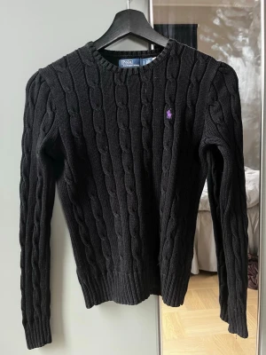  Polo Ralph Lauren tröja - Helt oanvänd Kabelstickad svart tröja från Polo Ralph Lauren i mjuk pima bomull. Klassisk rund halsringning, långa ärmar och lila broderad logga på bröstet. Perfekt för en stilren och tidlös look. 