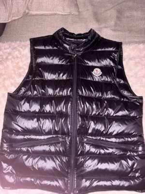 Svart dunväst från Moncler - Snygg svart dunväst från Moncler med glansig finish och klassisk logga på bröstet. Västen har hög krage, två dragkedjeförsedda fickor framtill och quiltad design. Perfekt för lager på lager och streetstyle.