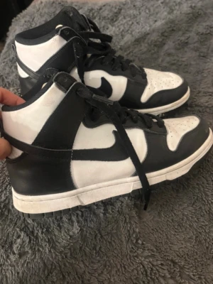 Nike Dunk High svartvita sneakers - Säljer ett par Nike Dunk High sneakers i svart och vitt läder med klassisk Swoosh på sidan. Skorna har högt skaft, svarta skosnören och perforerad tåbox för extra ventilation. Röd Nike-logga på plösen ger en snygg kontrast. Perfekta för dig som gillar retrostil.