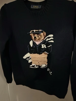 Svart stickad tröja Polo Ralph Lauren - Svart stickad tröja från Polo Ralph Lauren med ikonisk björn på bröstet. Tröjan har rund halsringning och långa ärmar. Björnen är broderad och bär collegekläder i blått, vitt och beige. Perfekt för dig som gillar klassisk preppy stil.