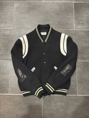 Saint Laurent Teddy varsity jacket - Saint Laurent Teddy varsity jacket in good condition.  The chain hanger has fallen off. The elbows have been professionally repaired with real leather patches. There is also some slight yellowing on the cuffs and waistband, but nothing major.  From 2018, still designed by Hedi Slimane.  Size: 48 (refer to measurements)  Measurements: Shoulders: 48 cm Pit to pit (chest): 55 cm Sleeve: 60 cm Length: 59 cm 