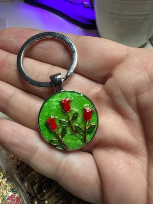 Nyckelring med röda rosor och grön bakgrund - Rund nyckelring i metall med en grön bakgrund och tre upphöjda röda rosor med gröna blad. Perfekt accessoar för dig som gillar färgglada detaljer och blommotiv.