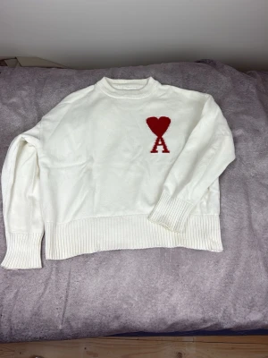 Vit stickad tröja från Ami - Säljer en stilren vit stickad tröja från Ami Alexandre Mattiussi med röd hjärtlogga och bokstaven A på bröstet. Tröjan har ribbade muddar vid ärmslut och nederkant samt rund hals. Perfekt för dig som gillar minimalistisk design.