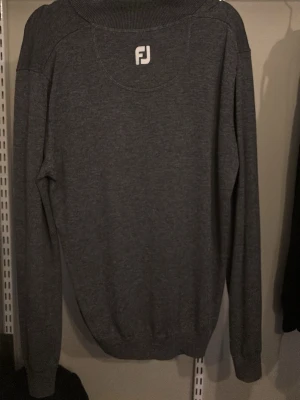 Mörkgrå stickad tröja från FootJoy - Mörkgrå stickad tröja från FootJoy med halv dragkedja och ribbad krage. Tröjan har långa ärmar och ribbade muddar vid ärmslut och nederkant. Perfekt för lager på lager och enkel att matcha med andra plagg. 