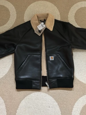 Svart skinnjacka från Carhartt - Snygg svart skinnjacka från Carhartt med beige fuskpäls på kragen och innerfodret. Jackan har två snedställda fickor fram, ribbade muddar och dragkedja. Klassisk Carhartt-logga på vänster ficka. Perfekt för dig som vill ha en stilren och trendig look. Oanvänd med lappen kvar.        Storlek M. Ny pris 3900 kr 