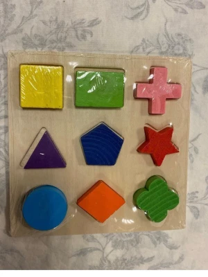 Träpussel med färgglada former - Ett träpussel med nio olika färgglada former, bland annat cirkel, triangel, stjärna och fyrklöver. Pusselbitarna är målade i blått, rött, grönt, gult, lila, orange och rosa. Perfekt för att lära sig former och färger. 3 år + 