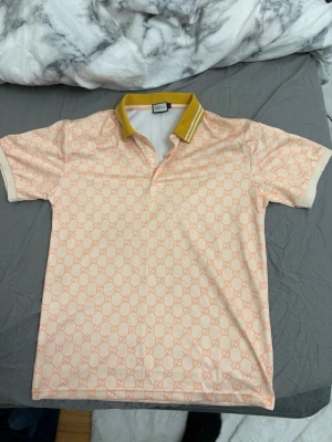 Gucci beige mönstrad polotopp - Säljer en beige polotopp från Gucci med ikoniskt GG-mönster i ljus orange över hela plagget. Kragen är gul med vita och orange detaljer. Klassisk pikéknäppning framtill och korta ärmar. Perfekt för dig som vill ha något unikt och trendigt. M passar S