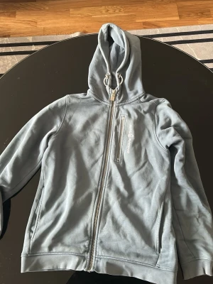 Ljusblå hoodie från sail racing - Ljusblå hoodie med dragkedja från sail racing. Tröjan har huva med dragsko, en bröstficka med dragkedja och två sidofickor. Tillverkad i mjukt material som känns skönt mot huden. Perfekt för chill dagar eller när du vill ha en avslappnad stil.