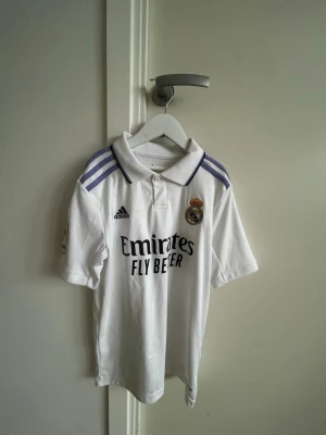 Real Madrid matchtröja Adidas vit - Säljer en vit Real Madrid tröja från 2023.matchtröja från Adidas med lila ränder på axlarna och klubbmärke på bröstet. Tröjan har korta ärmar, krage och trycket 'Emirates Fly Better' på framsidan. 