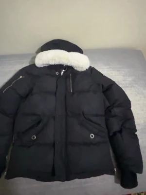 Svart dunjacka från Moose Knuckles - Säljer en svart dunjacka från Moose Knuckles med vit päls  på huvan och ikonisk metallogo på ärmen. Jackan har dragkedja, fickor med tryckknappar och en bröstficka med dragkedja. Perfekt för kalla vinterdagar och riktigt snygg streetstil.