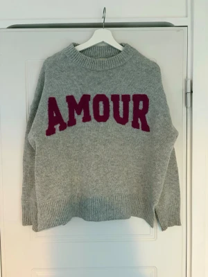 Grå stickad tröja med text AMOUR - Säljer en grå stickad tröja med stor vinröd text 'AMOUR' på bröstet. Tröjan har rund halsringning, ribbade muddar och avslut. Perfekt för dig som gillar en avslappnad och trendig look.
