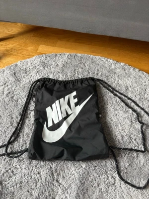 Svart Nike gympåse med logga - Cool svart gympåse från Nike med stor vit logga på framsidan. Väskan har snörstängning och tunna snören som axelband. Perfekt för träning eller när du vill ha. Med det finns det band till håret