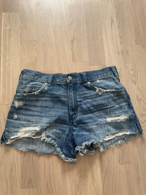 Blå slitna jeansshorts med fransar - Säljer ett par blå jeansshorts med slitna detaljer och fransig kant. Shortsen har fem fickor, bälteshällor och knappgylf. Perfekta för sommaren och ger en avslappnad, trendig look.