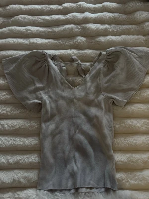 Vit ribbad topp med puffärm - Supersöt vit ribbad topp med stora puffärmar och v-ringning. Toppen har knyt i nacken och är figurnära i modellen. Perfekt för dig som gillar trendiga detaljer och vill sticka ut lite extra. Toppen ser grå ut på bilderna men är mer vit i verkligheten. Hittade inte den vita på hemsidan men är samma modell som sista bilderna