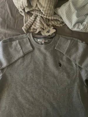 Grå sweatshirt från US Polo Assn - Snygg grå sweatshirt från US Polo Assn med rund halsringning och klassisk logga broderad på bröstet. Tröjan har långa ärmar och ribbade muddar vid ärmslut och nederkant. Perfekt för en avslappnad och stilren look. Aldrig använd! Med prislappen kvar. 