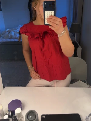 Röd cool blus - Säljer en röd blus med volangdetalj vid axlar och bröst. Blusen har en lös passform och är ärmlös, perfekt för varmare dagar. Enkel att styla med både jeans och kjol för en trendig look.