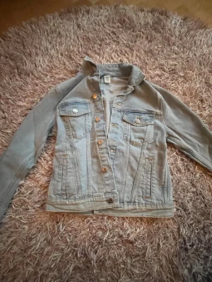 Ljusblå jeansjacka från H&M - Säljer en klassisk ljusblå jeansjacka från H&M i storlek 36. Jackan har två bröstfickor med lock och knappar, samt två sidofickor. Snygga sömdetaljer och metallknappar framtill. Perfekt att slänga över en hoodie eller t-shirt.