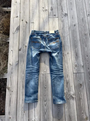 Vintage Nudie Jeans - Säljer ett par klassiska blå jeans från Nudie Jeans med snygga slitningar och kontrastsömmar. Modellen har raka ben och två bakfickor med den ikoniska Nudie-bågen i orange. Perfekta för en avslappnad och trendig look. Lagade på ena knät. Storlek W31 L32