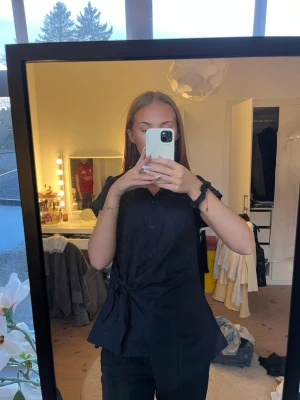 Mörkblå skjorttopp med knyt i midjan - Säljer en mörkblå skjorta med korta ärmar och klassisk krage. Skjortan har knappar framtill och ett snyggt knyt i midjan som ger en cool och modern look. Materialet känns som bomull och modellen är något lösare med asymmetrisk nederkant.