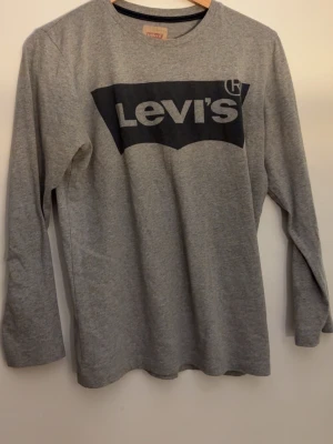 Grå långärmad topp från Levi's - Snygg grå långärmad topp från Levi's med stort svart logotryck på bröstet. Tillverkad i mjuk bomull och har en klassisk rund halsringning. Perfekt för en avslappnad och stilren look.