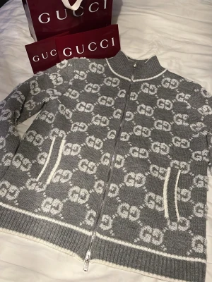Grå cardigan från Gucci - Säljer en grå stickad ziptröja från Gucci med vit GG-logga över hela tröjan. Tröjan har hög krage, hel dragkedja framtill och två fickor med vita detaljer. Ribbstickade muddar och nederkant. Perfekt för dig som gillar exklusiv streetstyle. Köpt från Gucci finns kvitto till.