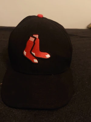 Boston Red Sox svart keps - Säljer en svart keps från New Era med Boston Red Sox-loggan broderad framtill i rött och vitt. Baksidan har MLB-loggan i vitt, svart och rött. Klassisk böjd skärm och röd knapp på toppen. Perfekt för dig som gillar baseball och streetwear.