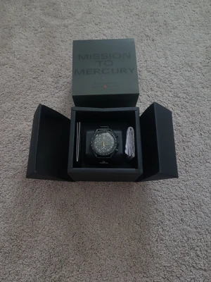 Omega x Swatch Mission to Mercury X Moon Remake - Snygg svart Omega x Swatch Speedmaster MoonSwatch Mission to Mercury X Moon Remake med svart boett och svart textilband. Klockan har vita detaljer på urtavlan och en gul sekundvisare. Kommer i originalbox med manual.