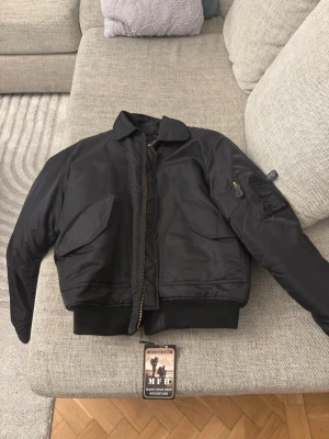 Svart bomberjacka från MFH - Klassisk svart bomberjacka från MFH, inspirerad av US Airforce CWU-45/P. Jackan har två stora fickor framtill, ärmficka med dragkedja och ribbade muddar. Materialet är slitstark nylon med polyesterfoder. Perfekt för dig som gillar en tidlös och cool look. Kom gärna med prisförslag!