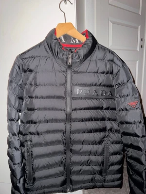 Svart dunjacka från Prada - Snygg svart dunjacka från Prada med diskret logga på bröstet och trekantsmärke på ärmen. Jackan har quiltad design, dragkedja framtill och praktiska fickor. Insidan har en röd detaljficka med dragkedja. Perfekt för kalla vinterdagar.